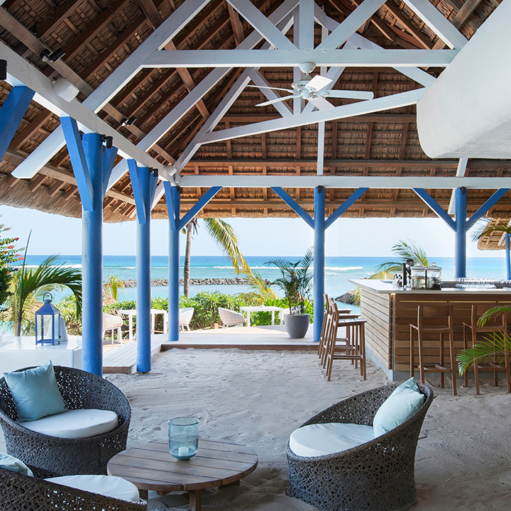 Beach bar at Veranda Pointe aux Biches with blue columns, thatched roof, woven lounge chairs, and ocean view beyond palm trees.