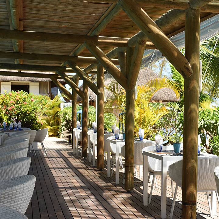Wooden pergola shades a dining terrace at Veranda Pointe aux Biches with white chairs, tables, and tropical plants in the background.