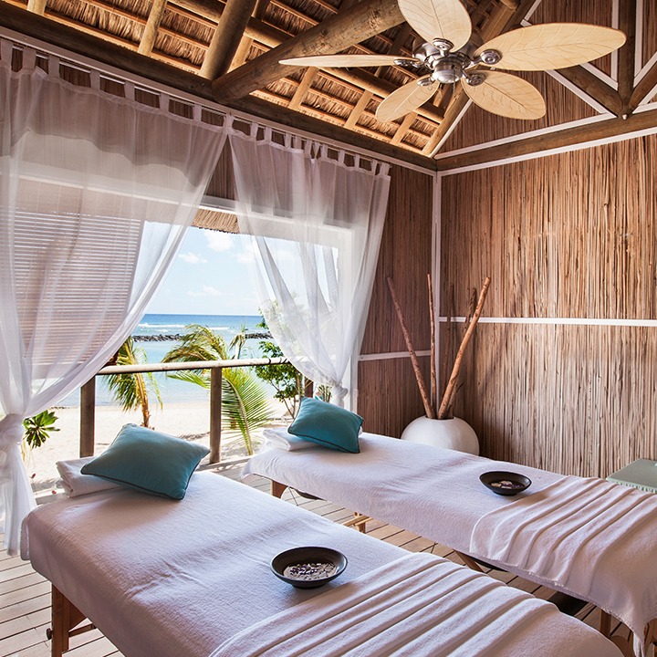 Twin beds with teal pillows in a thatched bungalow at Veranda Pointe aux Biches, ocean and palm trees visible through sheer curtains.