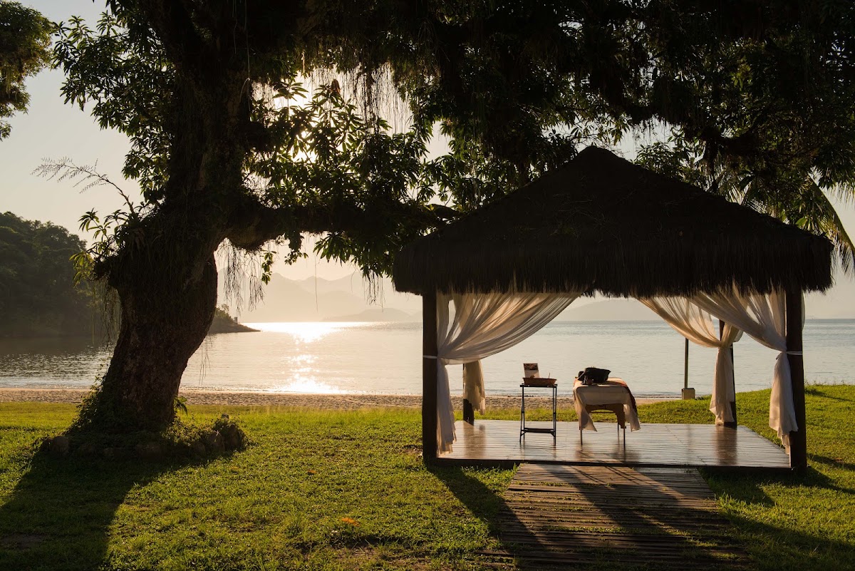 A person relaxes under a thatched pavilion with sheer curtains on the waterfront at Vila Galé Eco Resort de Angra, with a large tree overhead and calm bay waters beyond.
