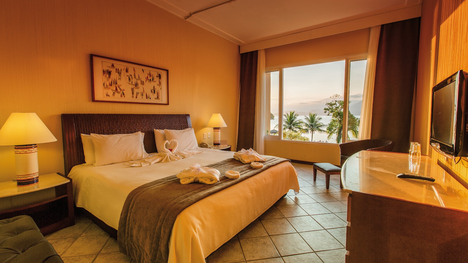 Warm-toned resort bedroom with king bed, yellow and brown bedding, ocean view through glass doors, palm trees visible outside, and desk area with TV.