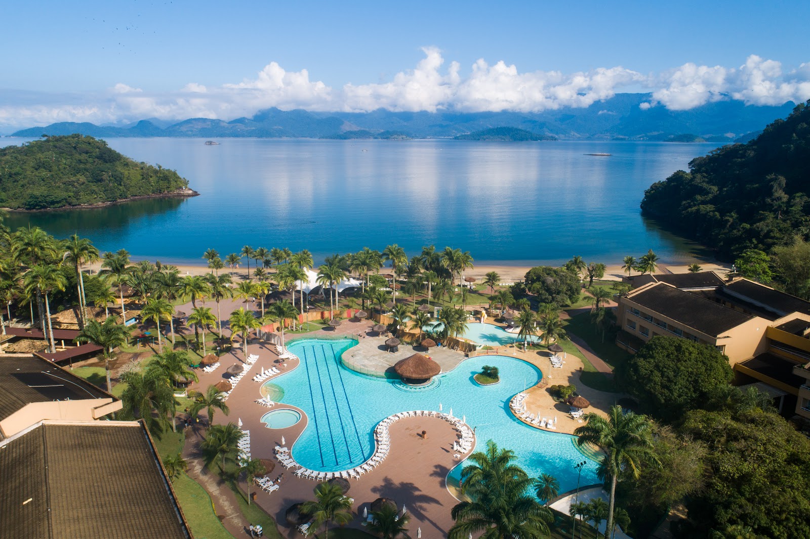 Vila Galé Eco Resort de Angra's curved pool complex with palm trees overlooks a sandy beach and blue bay surrounded by forested mountains.