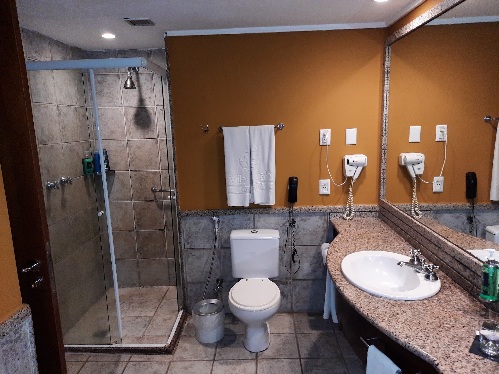 Bathroom with glass shower enclosure, toilet, and dual sink vanity with granite countertop against orange walls and gray tile accents.