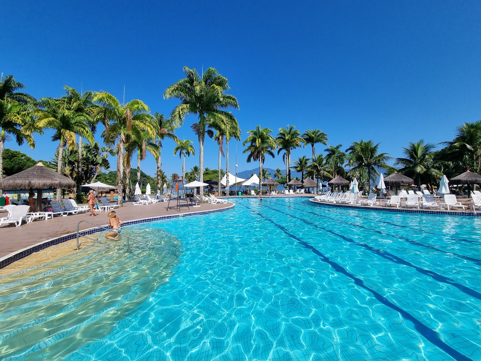 Clear blue swimming pool with lap lanes at Vila Galé Eco Resort de Angra, surrounded by palm trees, white loungers, and thatched umbrellas under a cloudless sky.