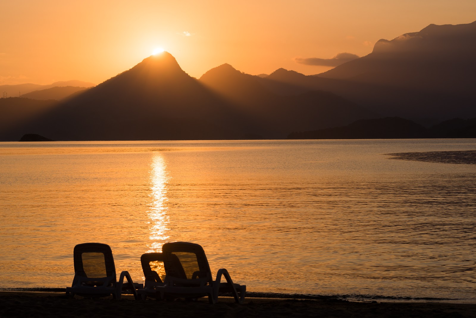 Two lounge chairs on a beach at sunset, with mountains silhouetted against an orange sky and calm water reflecting sunlight.