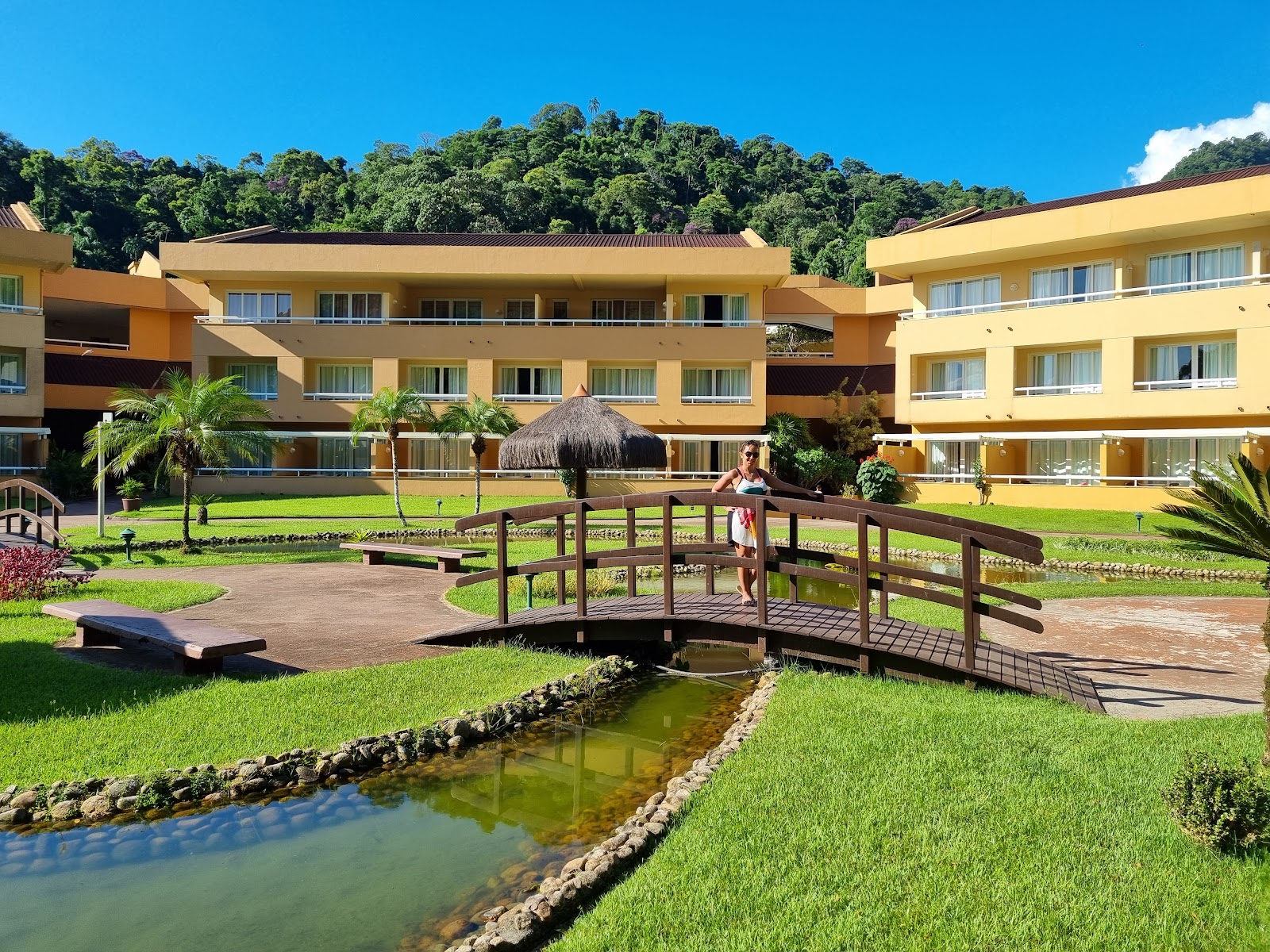 Yellow resort buildings with manicured lawns, a wooden footbridge crossing a small pond, palm trees, and forested hills under a blue sky.