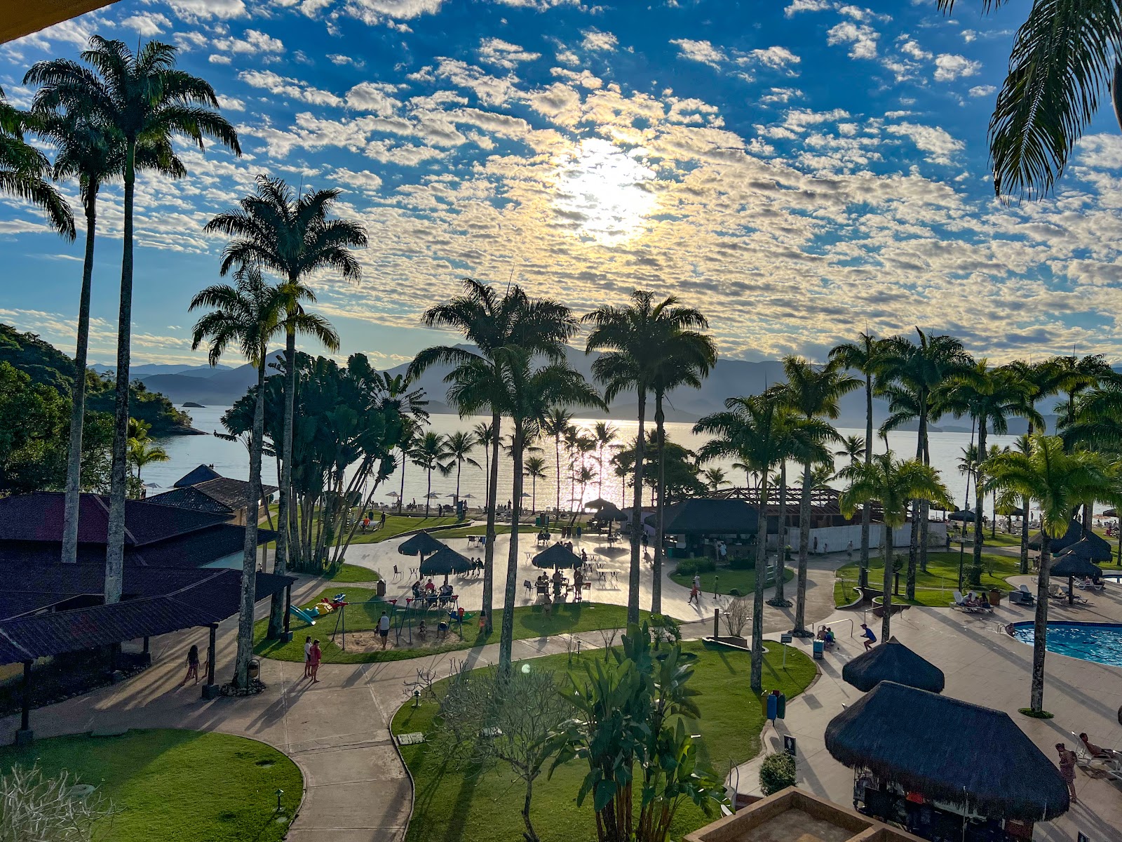 Vila Galé Eco Resort de Angra's grounds feature palm trees, manicured lawns, thatched-roof structures, and a view of the bay under a partly cloudy sky.