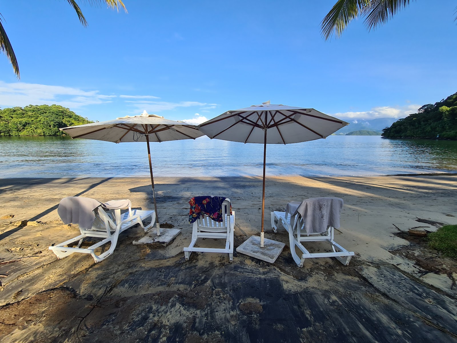 Sandy beach with shallow turquoise water, dense tropical vegetation, and mountains across the bay under blue sky with white clouds.