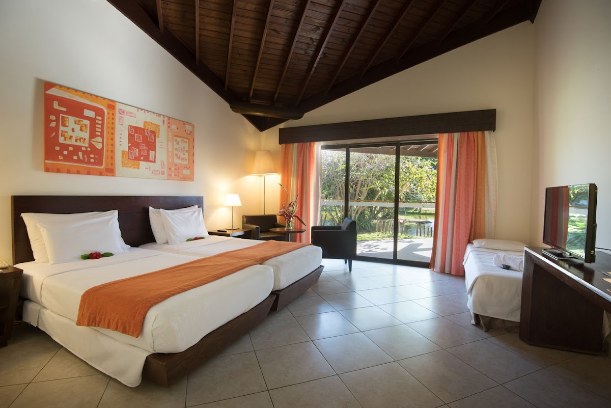 Resort bedroom with king bed, orange accent runner, dark wood ceiling, and sliding glass doors opening to garden grounds.