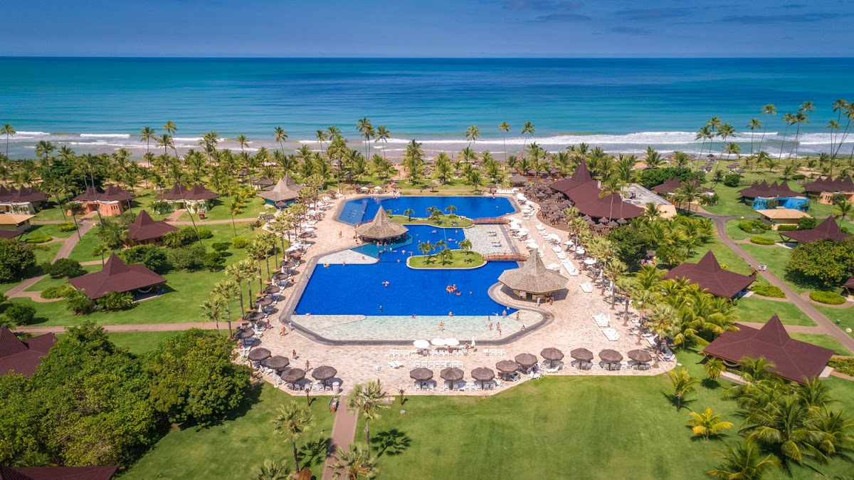 Aerial view of Vila Galé Marés with large blue pool complex, thatched umbrellas, palm-lined grounds, and ocean beach beyond.