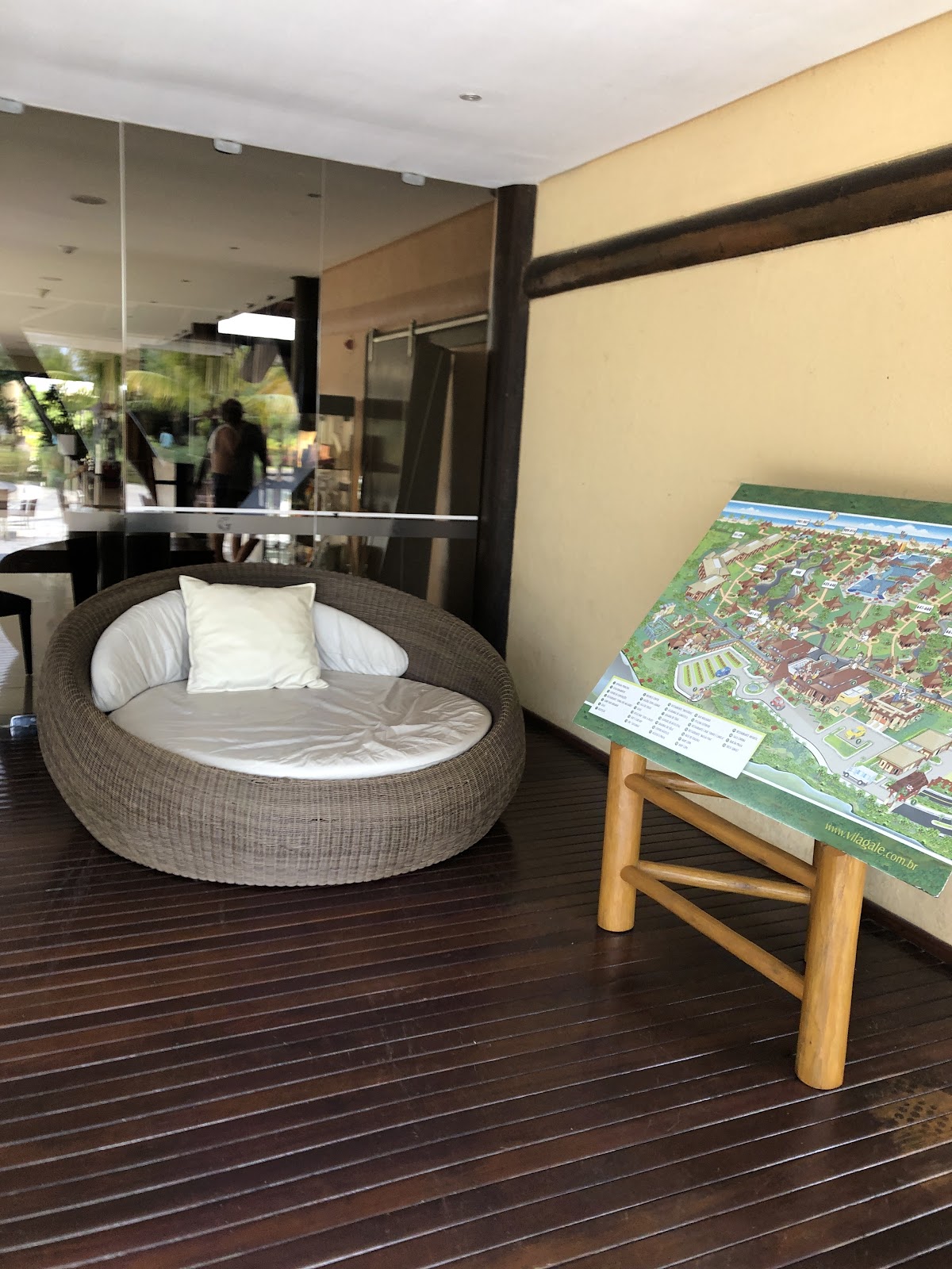 Round woven lounge chair with white cushions on dark wood deck, resort map on wooden stand, glass doors and yellow walls visible.