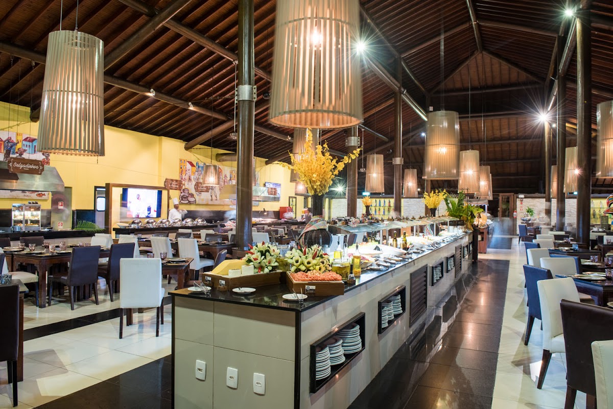Open-concept dining hall with central food stations, cylindrical pendant lights, and wooden beam ceiling at Vila Galé Marés.