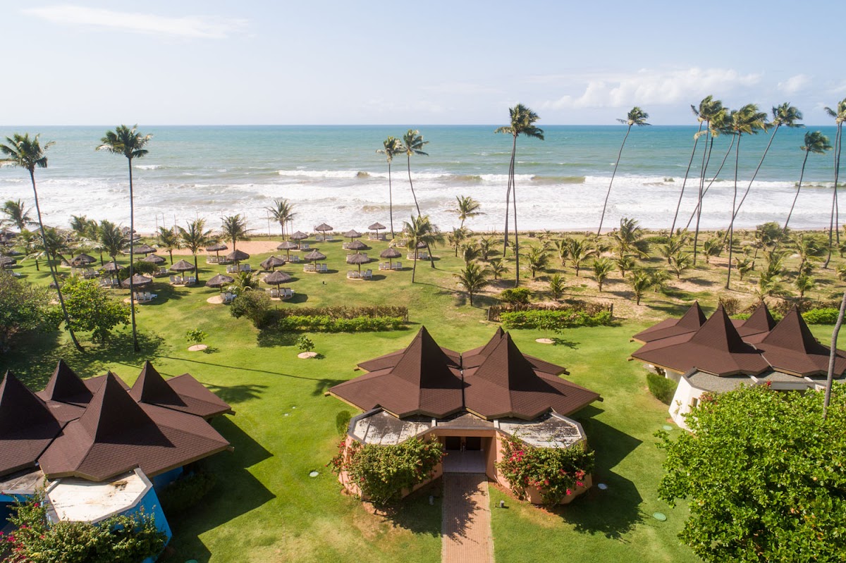 Aerial view of Vila Galé Marés beachfront with brown tent-shaped bungalows on manicured grass, thatched umbrellas, and palm trees overlooking the ocean.