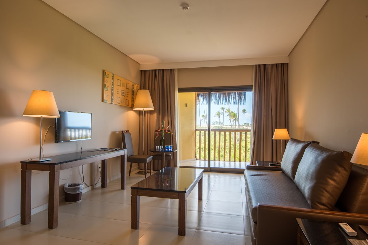 Resort suite with sofa, desk, and balcony overlooking palm trees and ocean at Vila Galé Marés.