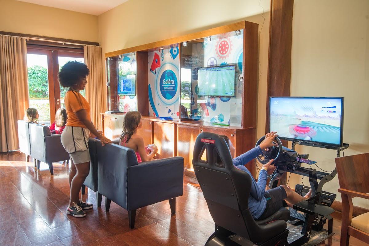 Kids' game room at Vila Galé Marés with racing simulator, arcade display, and children playing near floor-to-ceiling windows overlooking grounds.