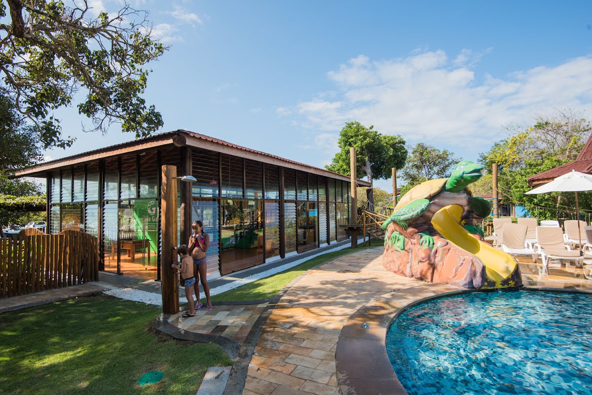 Modern wooden building with louvered windows next to a curved pool with a colorful slide, surrounded by lawn and lounge chairs.