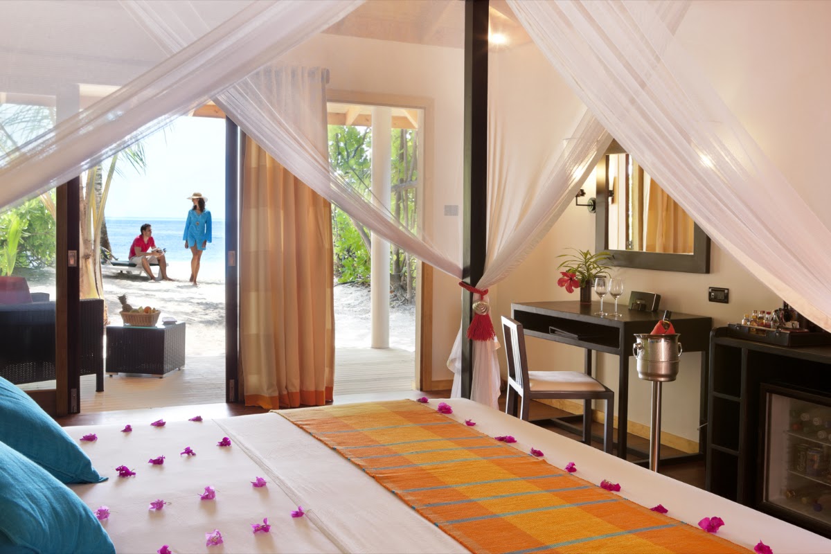 Resort villa bedroom with sheer white curtains, orange runner on bed scattered with pink flowers, ocean view through open doors to beach where two guests walk.