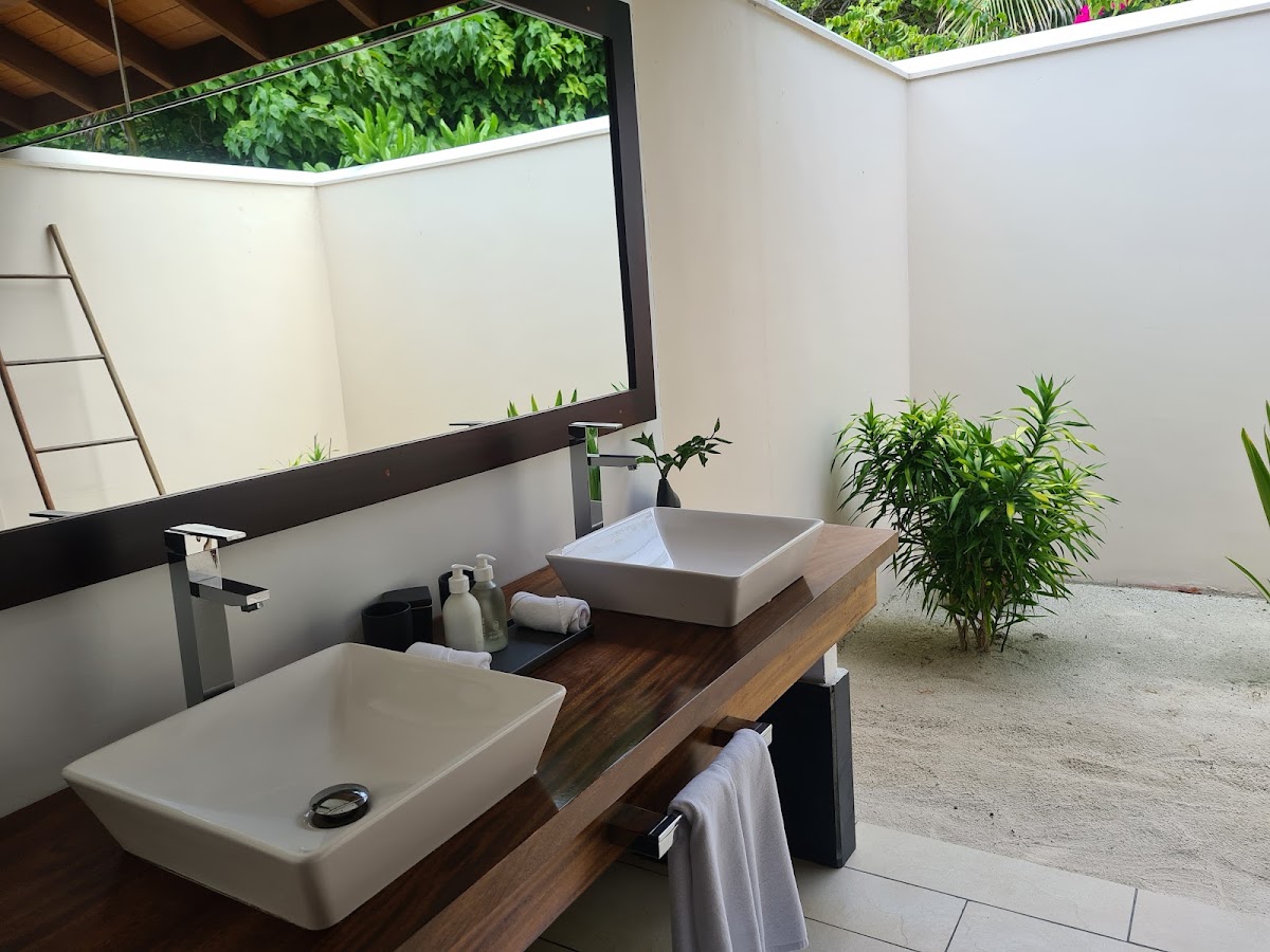 Open-air bathroom with two white vessel sinks on a wooden counter, large mirror, potted plants, and views of tropical greenery above.