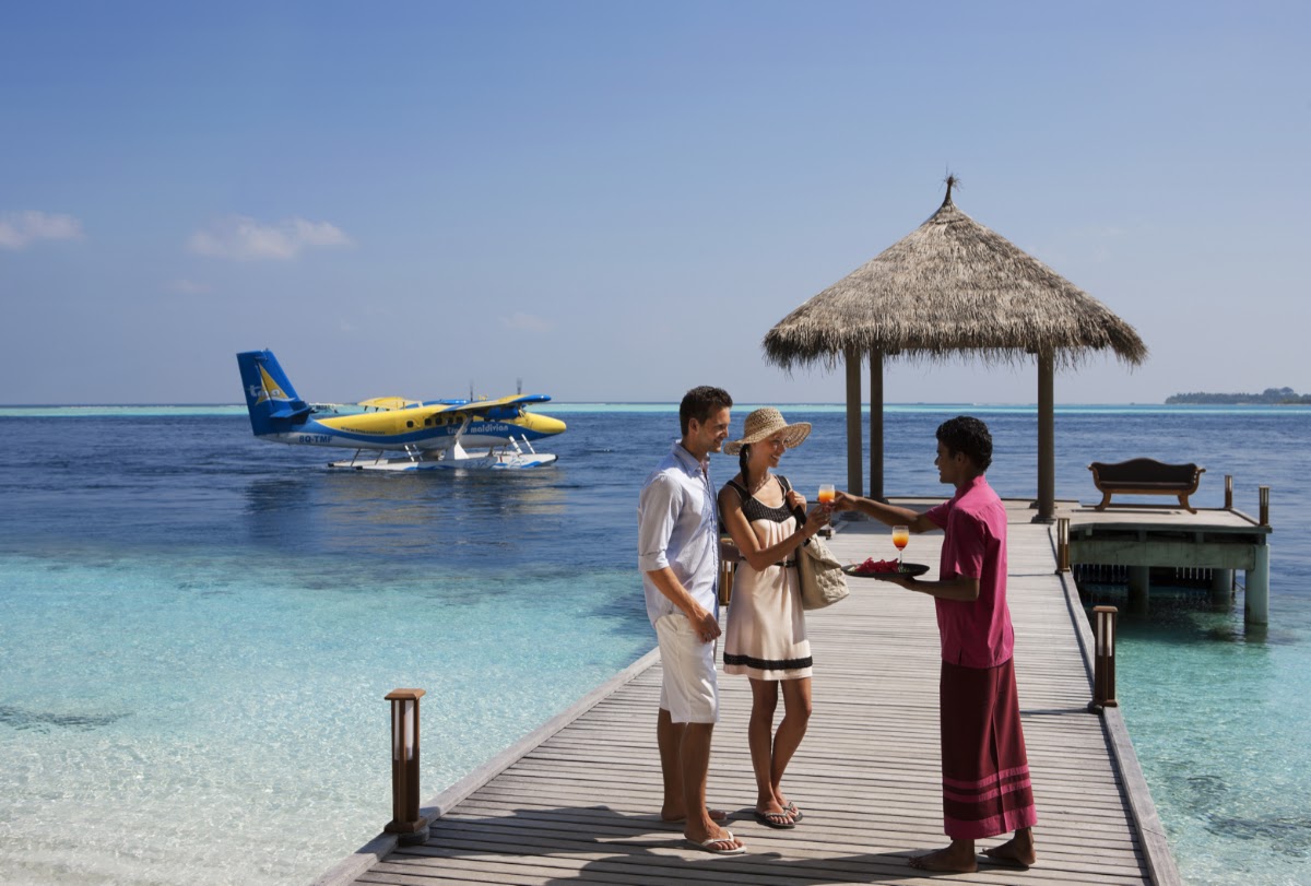 Couple on wooden pier receiving drinks from server, with seaplane and thatched-roof cabana in turquoise lagoon behind them.