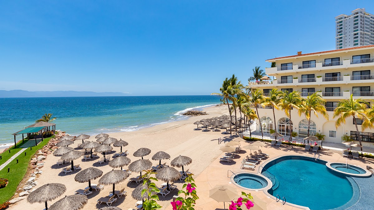 Villa del Palmar Beach Resort & Spa overlooks a sandy beach with thatched umbrellas, lounge chairs, and a curved pool under clear blue sky.