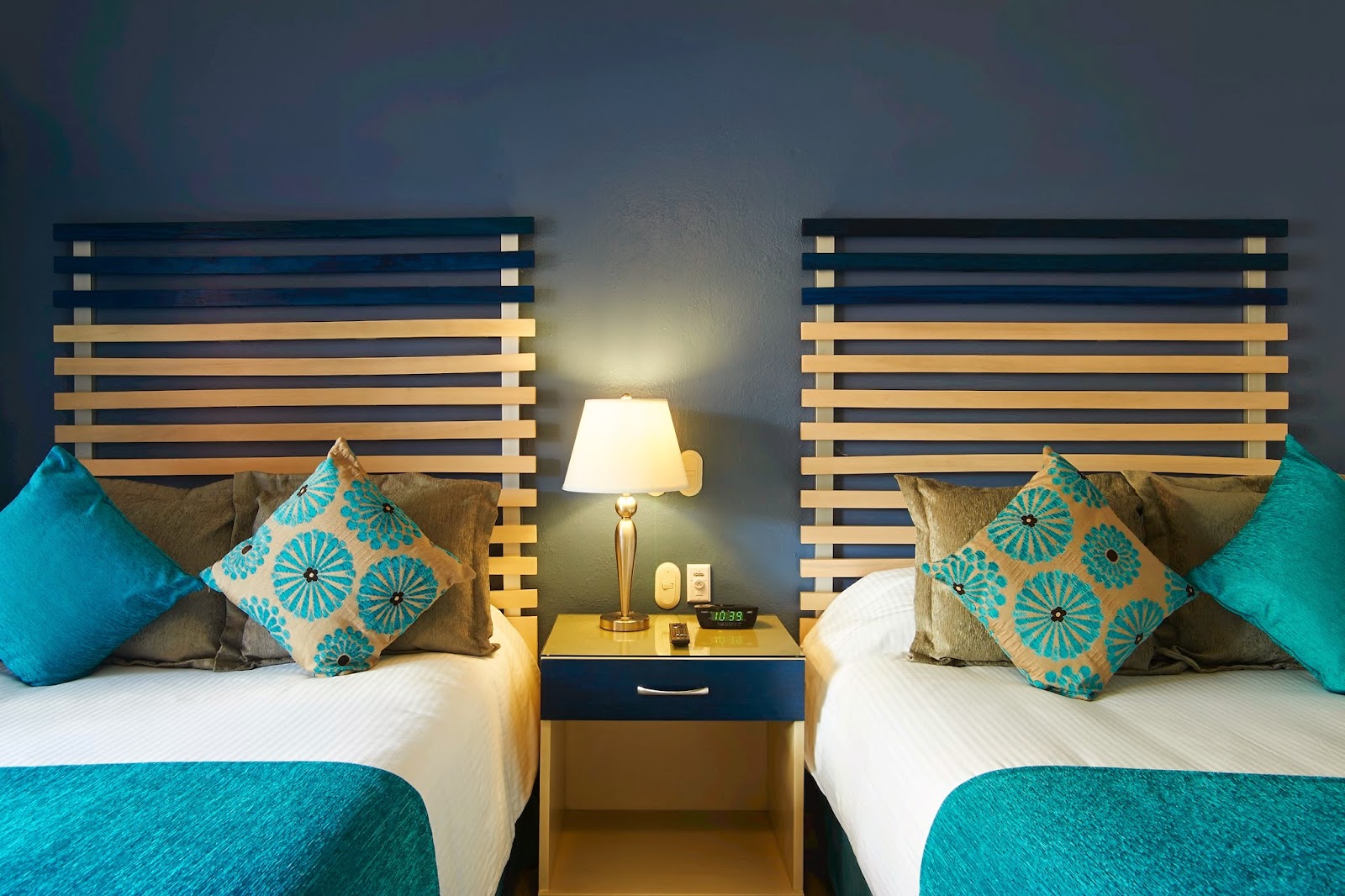 Twin beds with turquoise and patterned pillows separated by a nightstand with lamp in a modern resort bedroom.
