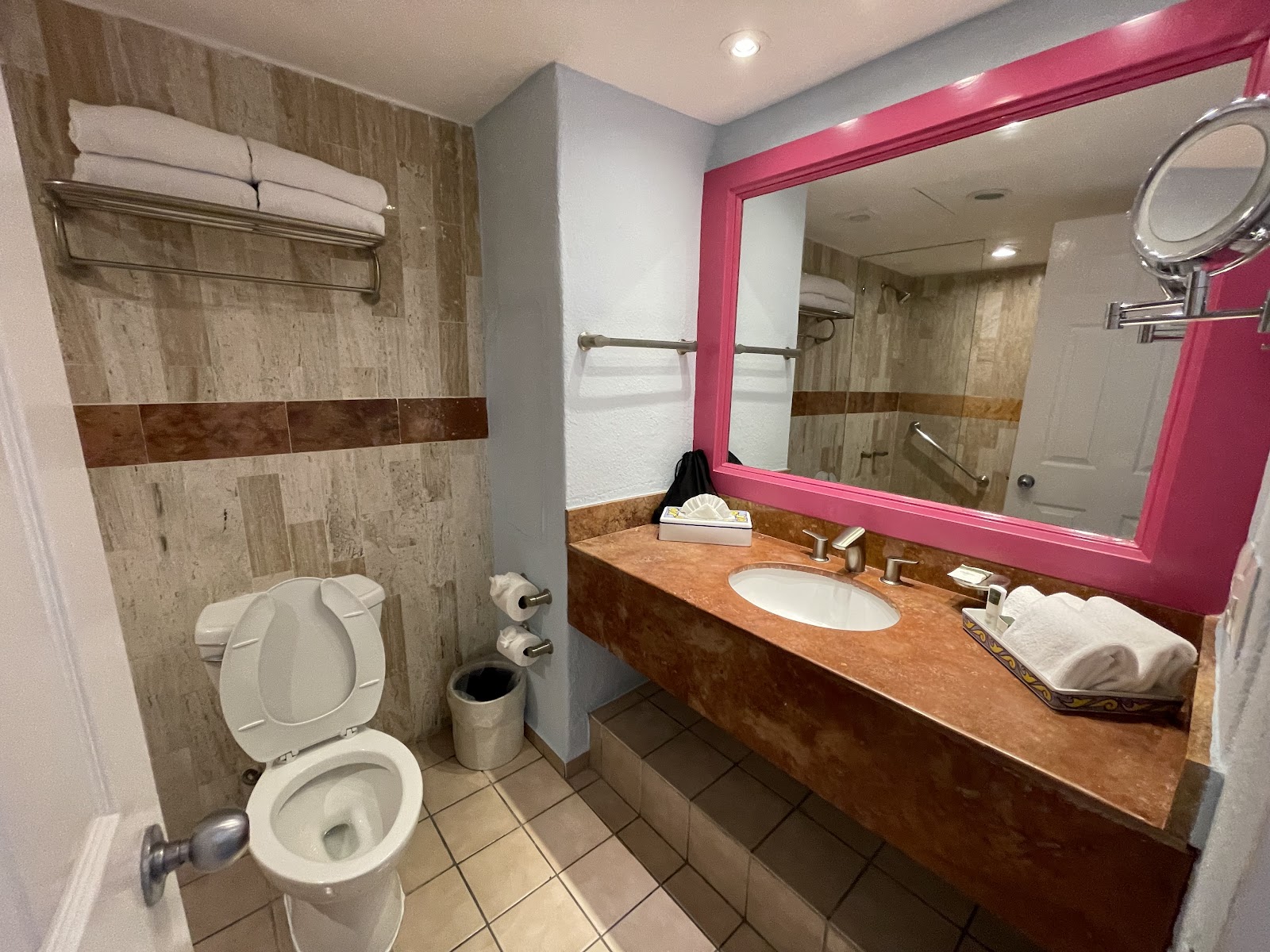 Resort bathroom with walk-in shower, brown vanity countertop, pink-framed mirror, and toilet with folded white towels on shelf.
