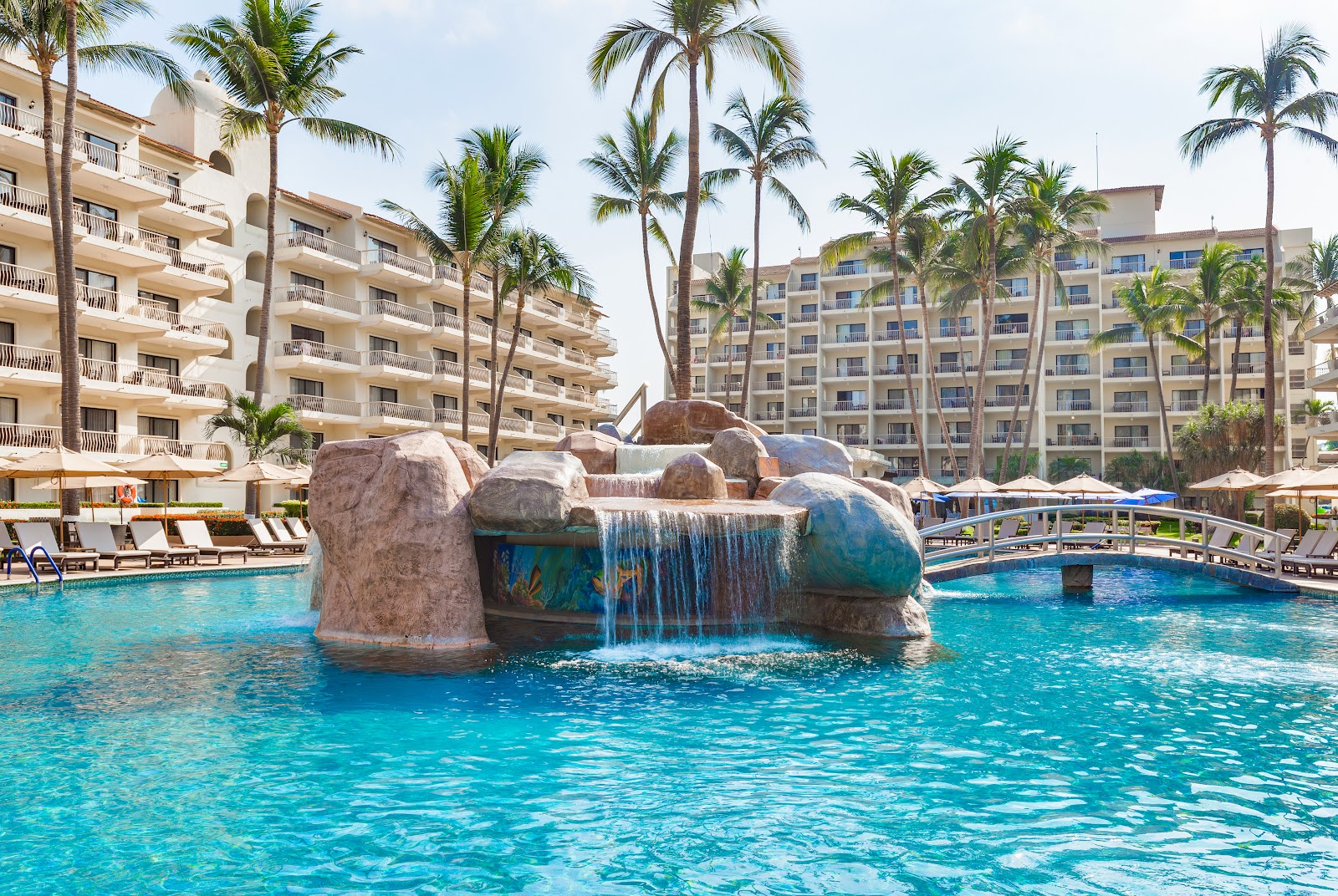 Large rock formation with waterfall in turquoise pool at Villa del Palmar, flanked by multi-story resort buildings and palm trees.