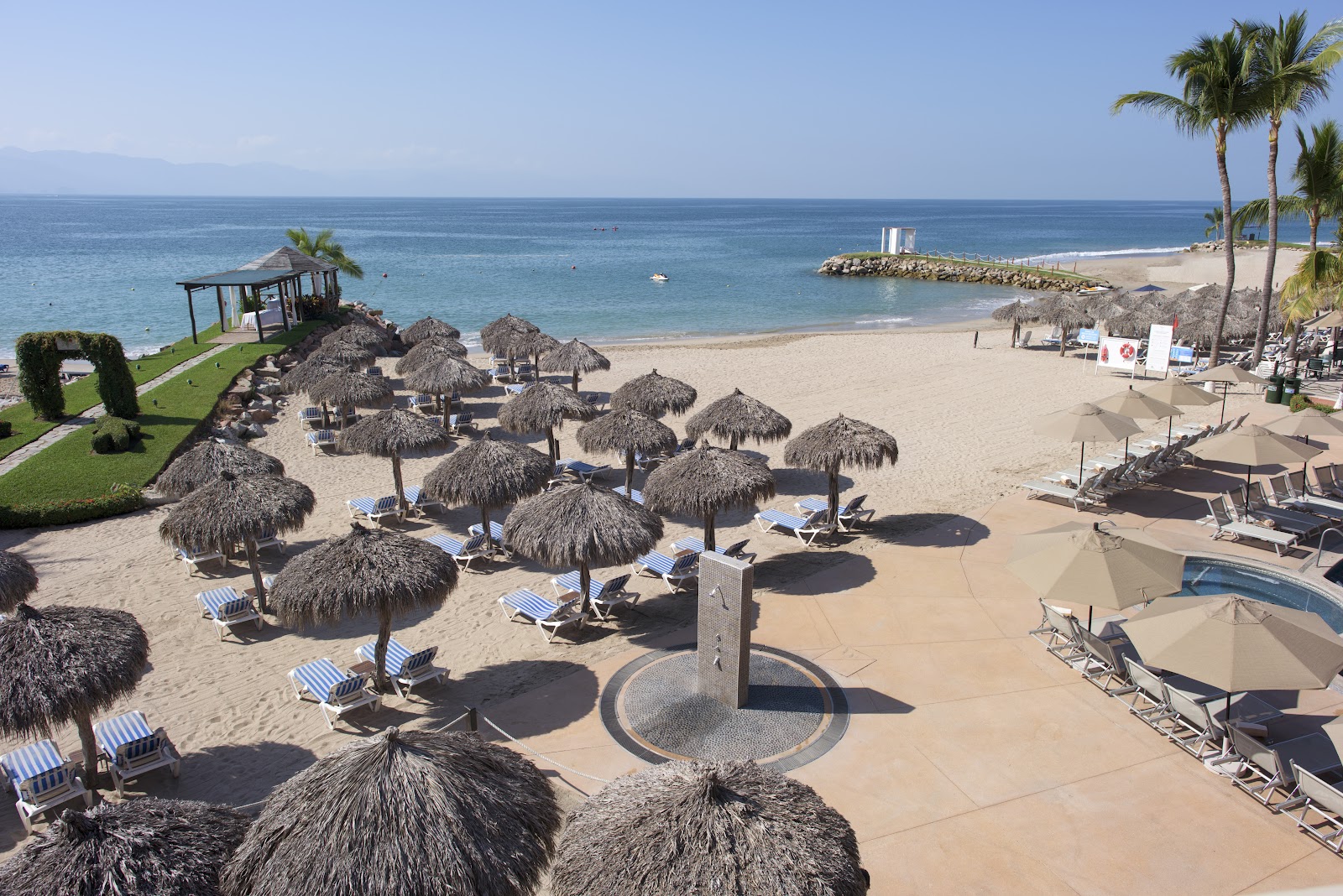 Resort pool with hot tub in foreground, lounge chairs and palapa umbrellas lining the deck, palm trees and beach beyond under clear blue sky.