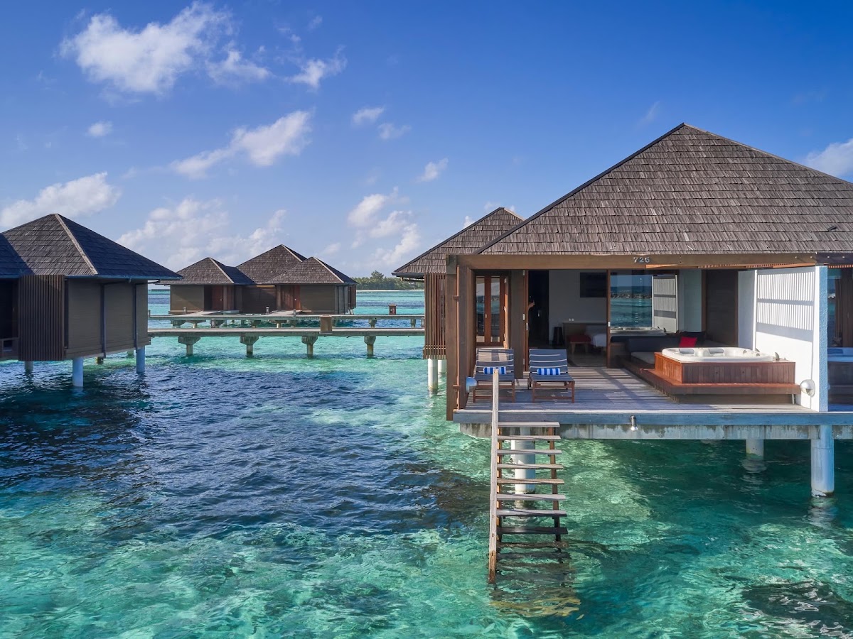 Overwater bungalows with peaked thatched roofs line turquoise lagoon waters at Villa Nautica, each with wooden decks and hot tubs.
