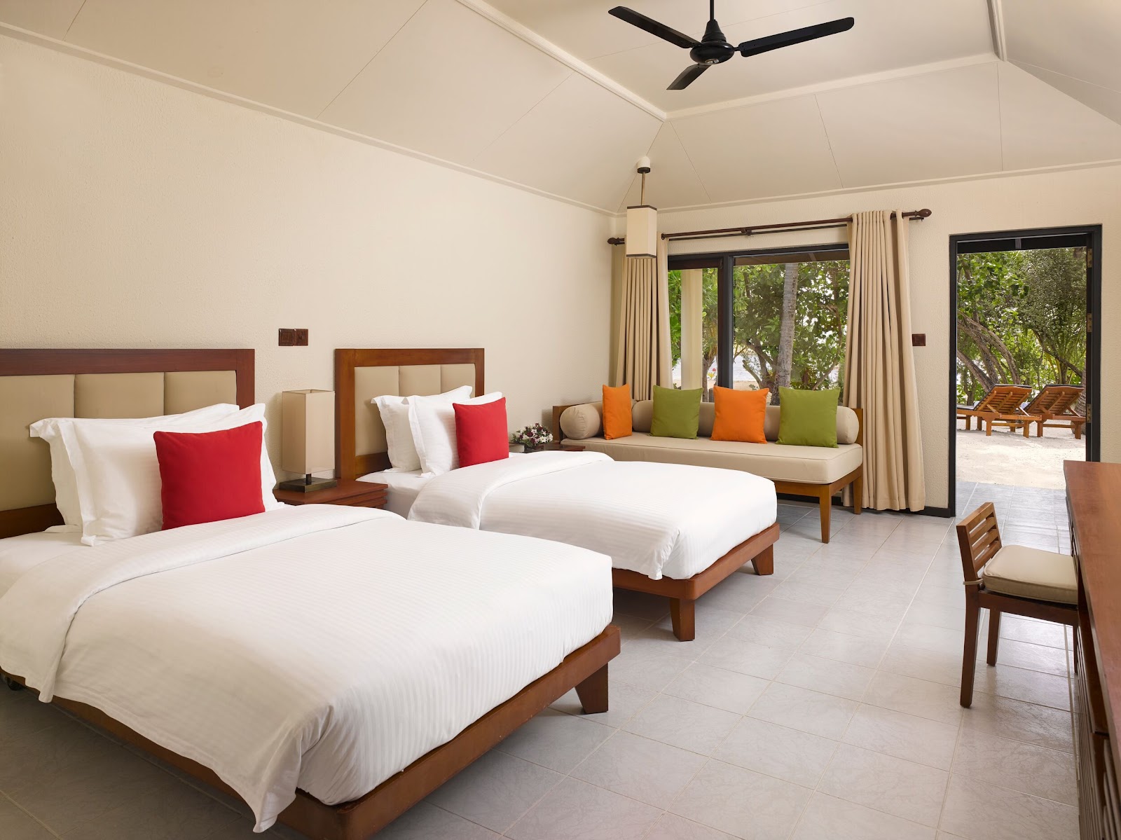 Spacious resort room with two queen beds, seating area with colorful pillows, and glass doors opening to a tropical deck with lounge chairs.