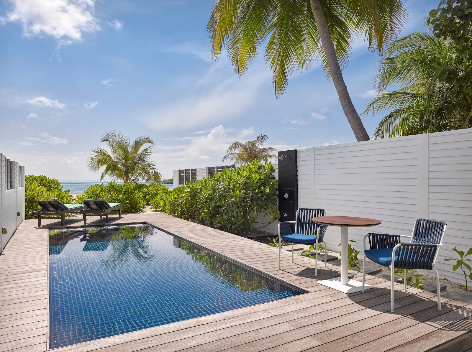 Private villa pool deck with blue tile pool, wooden loungers, dining table with chairs, white walls, palm trees, and ocean view beyond.