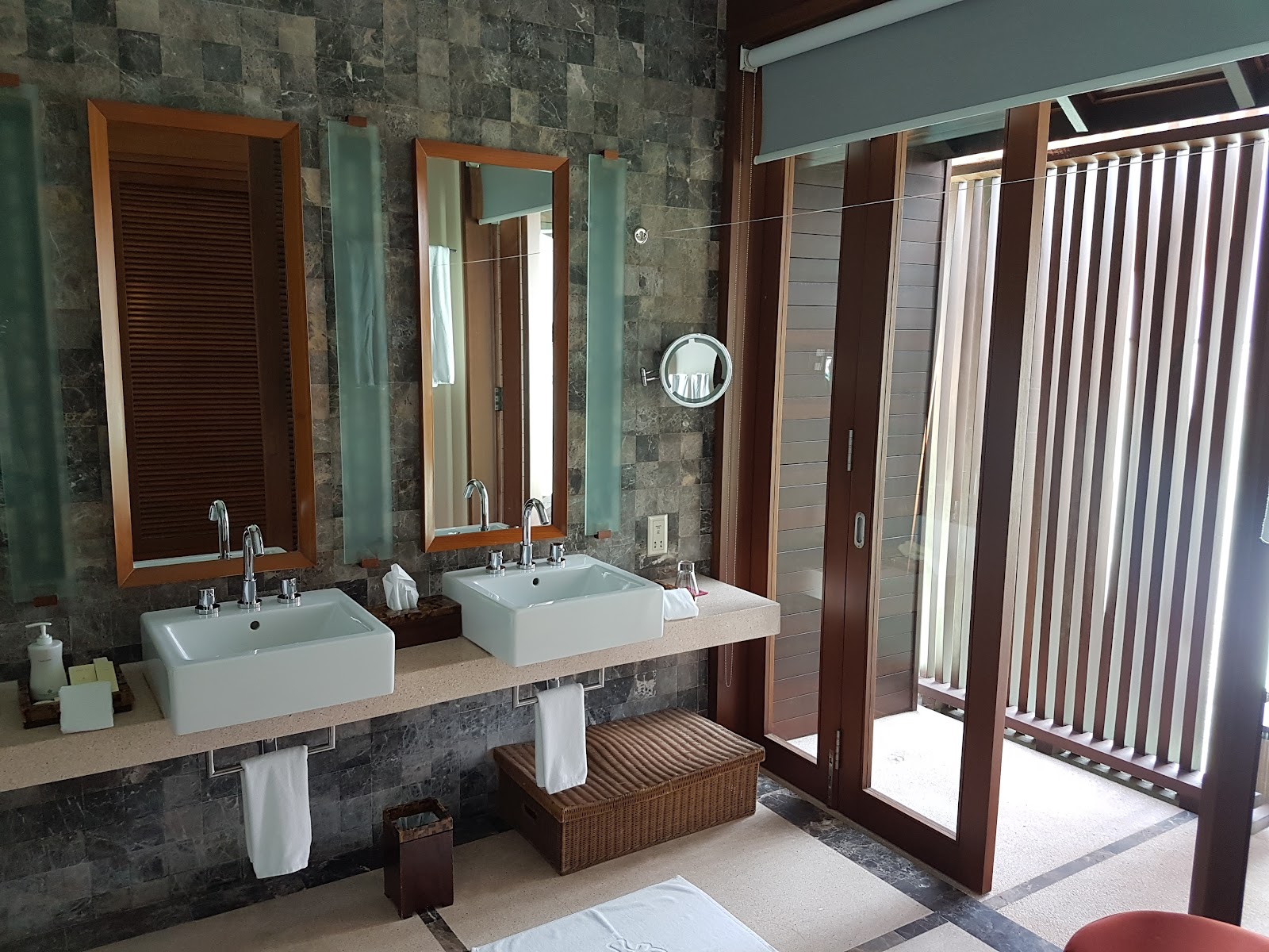 Bathroom with dual white vessel sinks, slate tile walls, wooden-framed mirrors, and glass doors opening to an outdoor shower area.