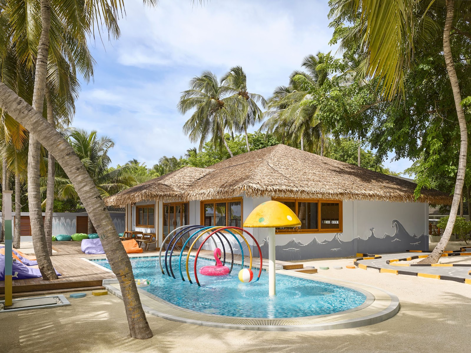 Shallow kids pool with colorful metal play structure and pink flamingo float, framed by palm trees and a thatched-roof bungalow at Villa Nautica.