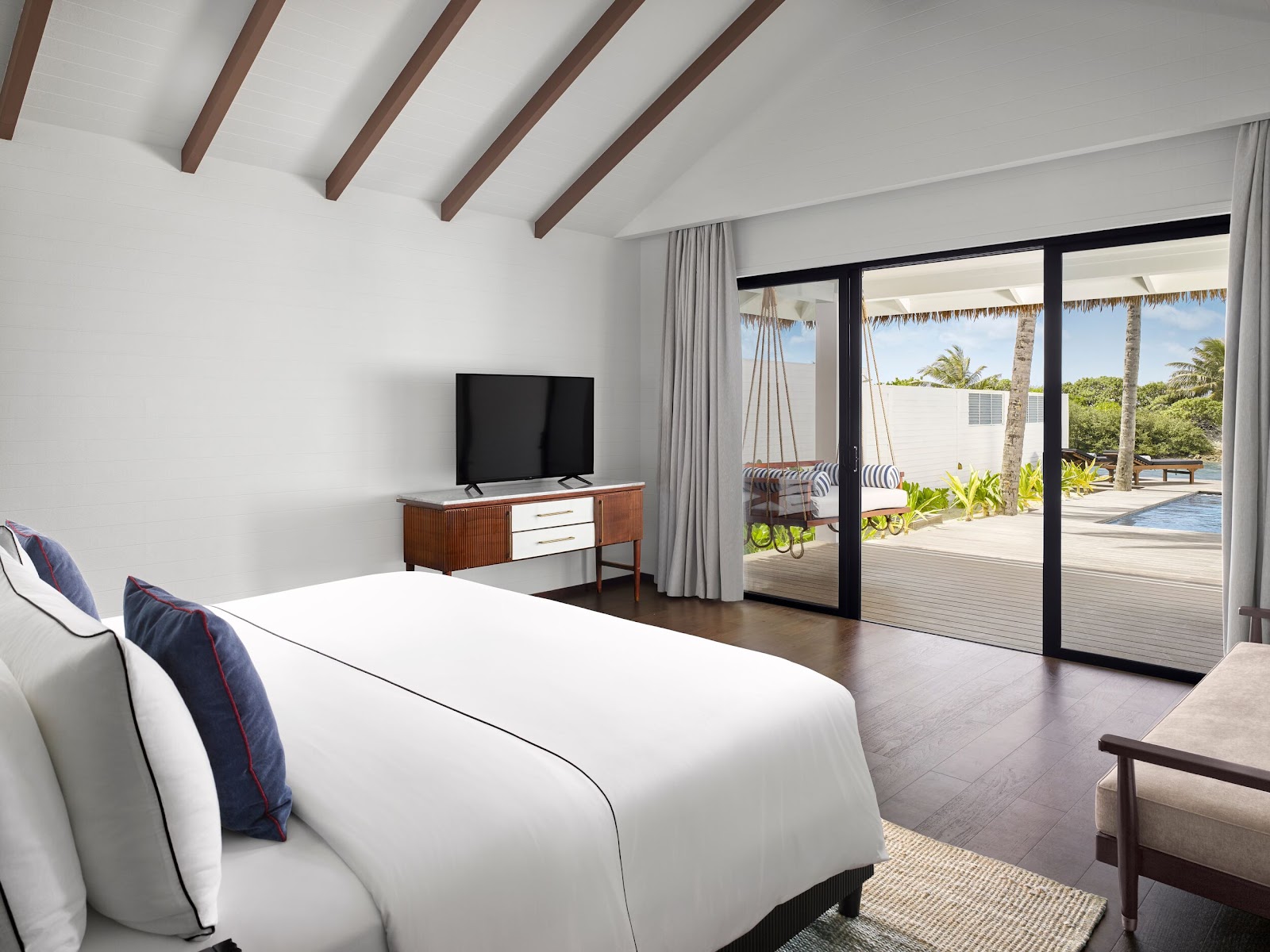 Resort bedroom with white bed, wooden beams overhead, and glass doors opening to a deck with striped loungers, palm trees, and ocean view.