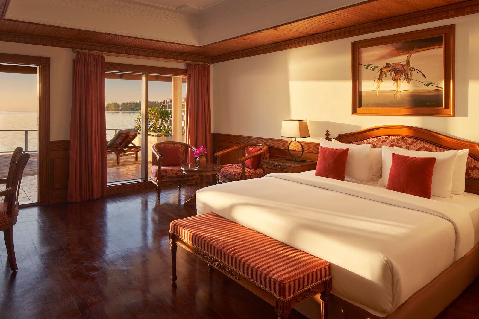 Spacious resort suite with wooden furnishings, a bed with red and white pillows, and glass doors opening to a waterfront balcony with lounge chairs.