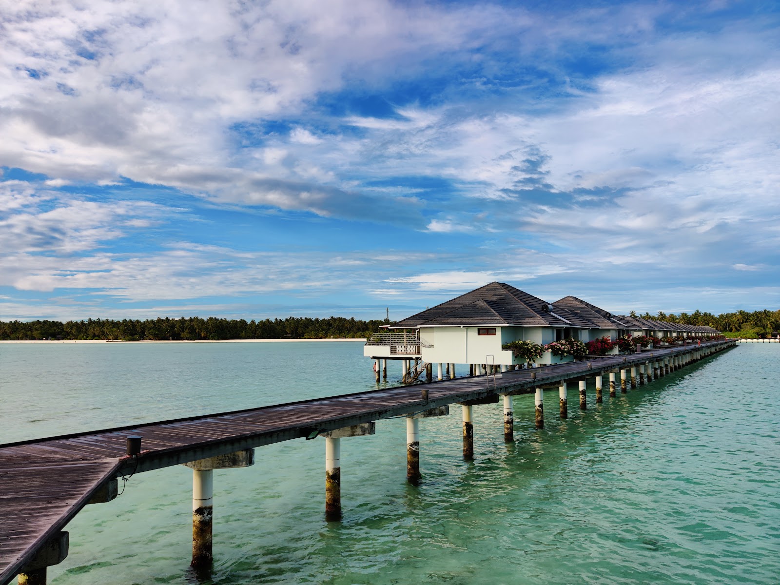 Aerial view of overwater bungalows and a central conical-roofed building at Villa Park, Maldives, with palm-lined island and turquoise lagoon.