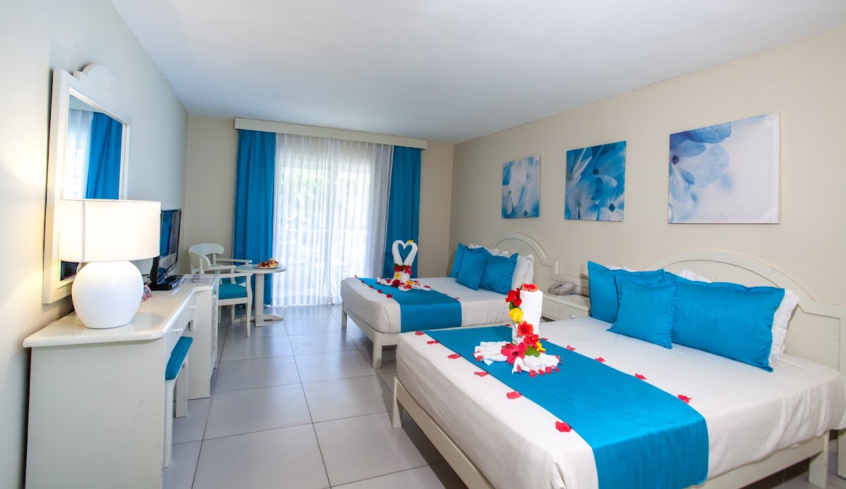 Resort guest room with two beds dressed in blue linens and red flower petals, blue curtains, and a desk area with lamp.