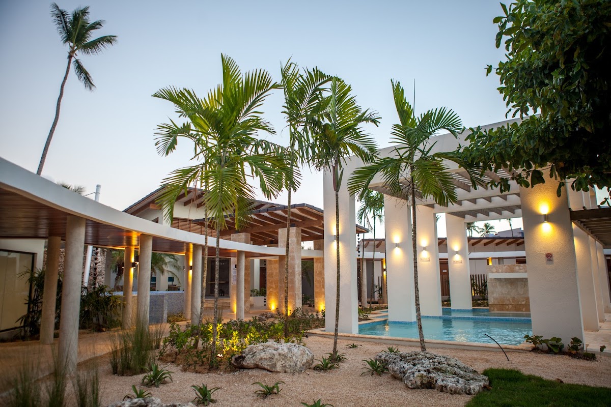 Vista Sol Punta Cana's courtyard at dusk features white buildings with wooden pergolas, palm trees, lit ground lights, and a blue pool reflecting the evening sky.