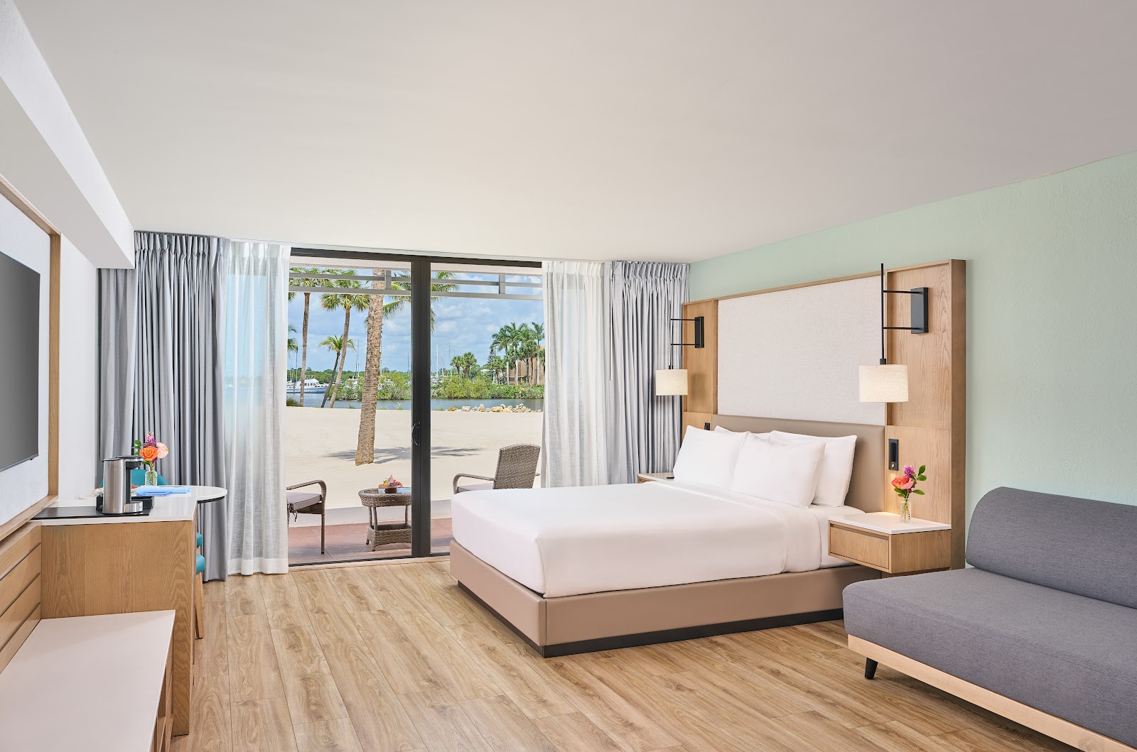 Modern beachfront resort bedroom with bed, sofa, and sliding glass doors opening onto sand and palm trees.