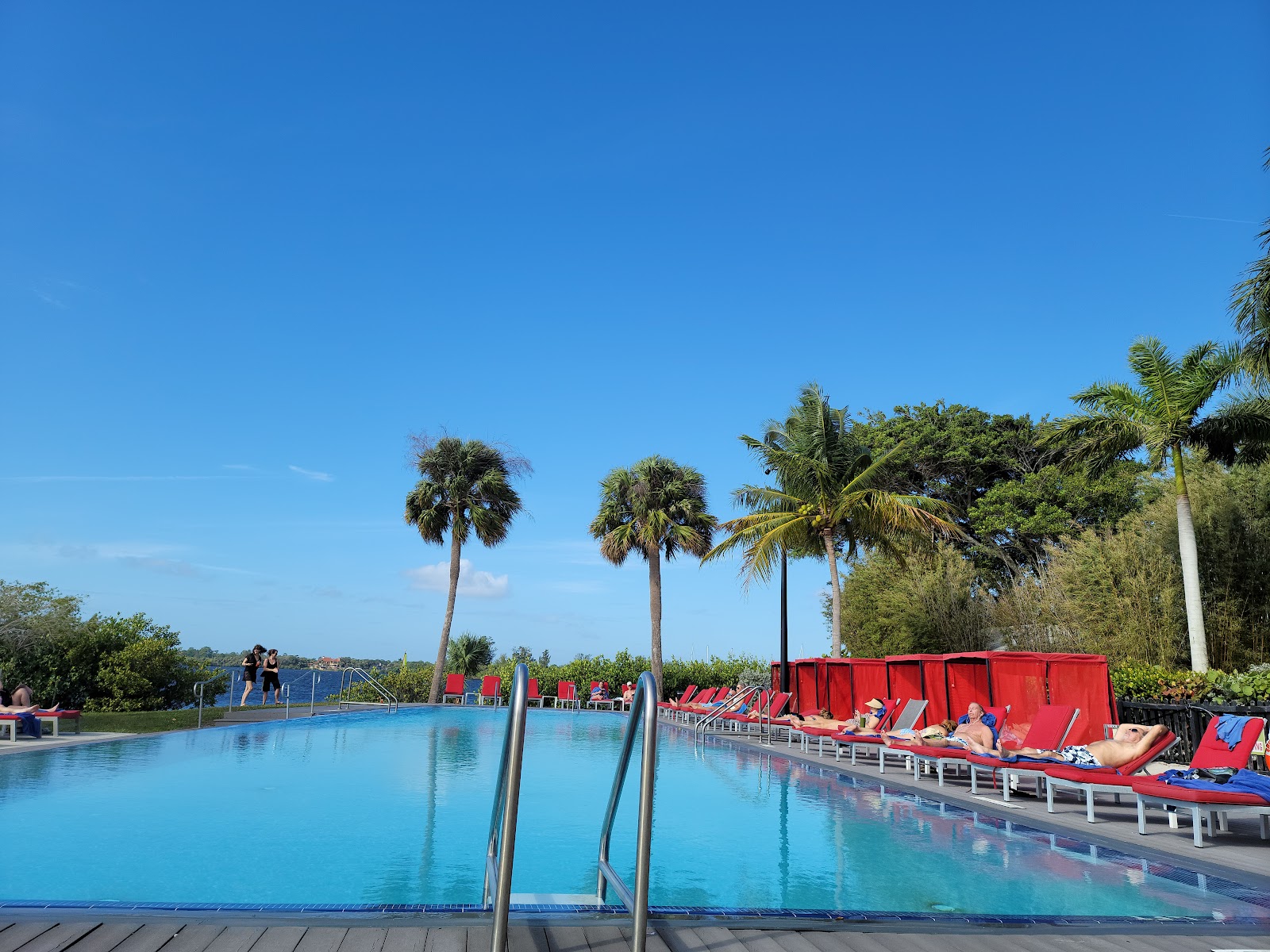Blue swimming pool with red lounge chairs lining the deck, palm trees, and guests relaxing under clear sky at voco Sandpiper All-Inclusive Resort.