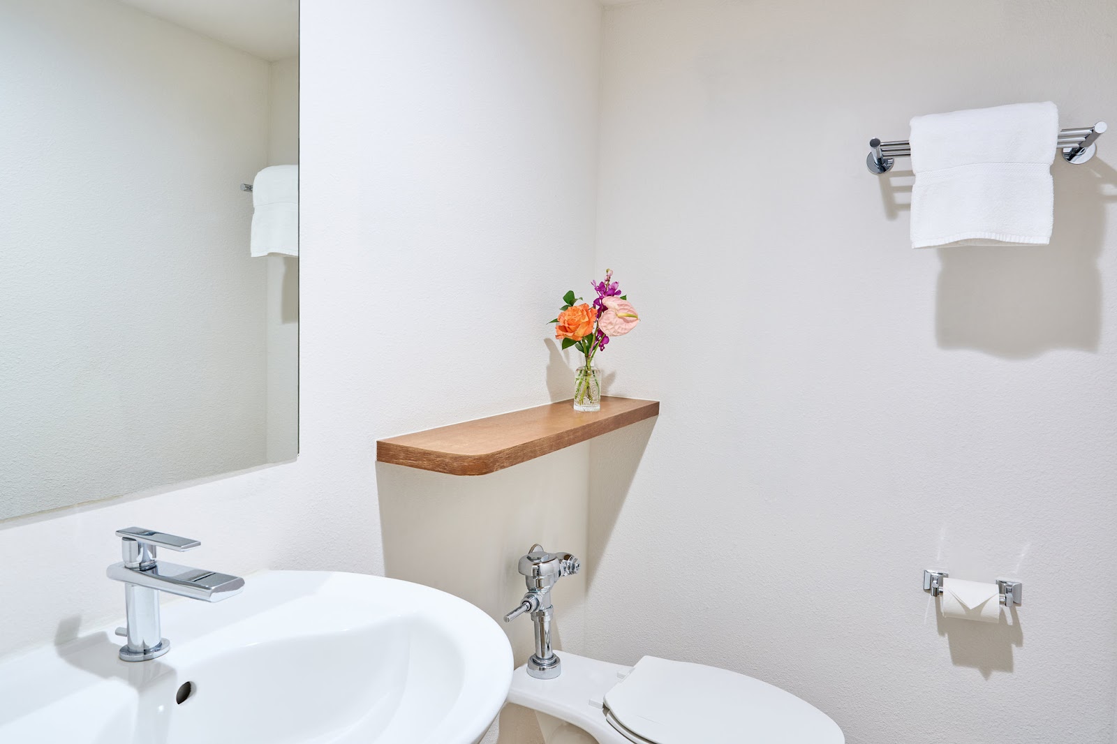 Clean white bathroom with round sink, chrome faucet, and a wooden shelf holding a vase of orange and pink flowers above the toilet.