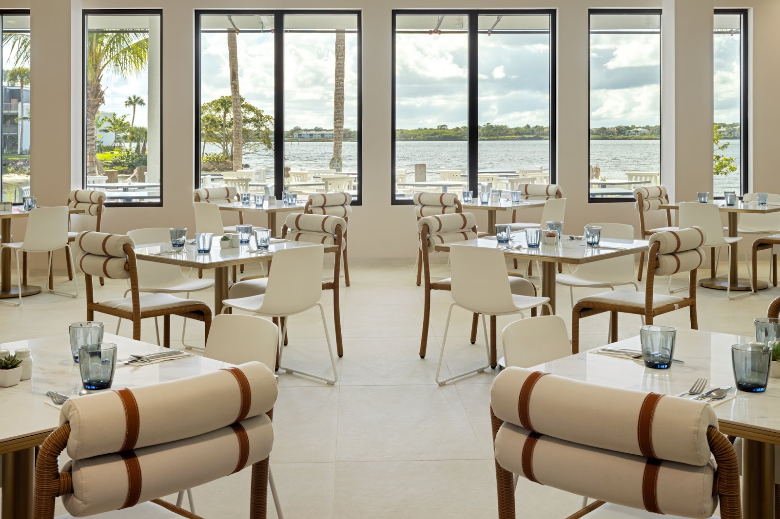 Modern dining room with beige chairs and white tables overlooking a waterway with palm trees and buildings visible across the water.