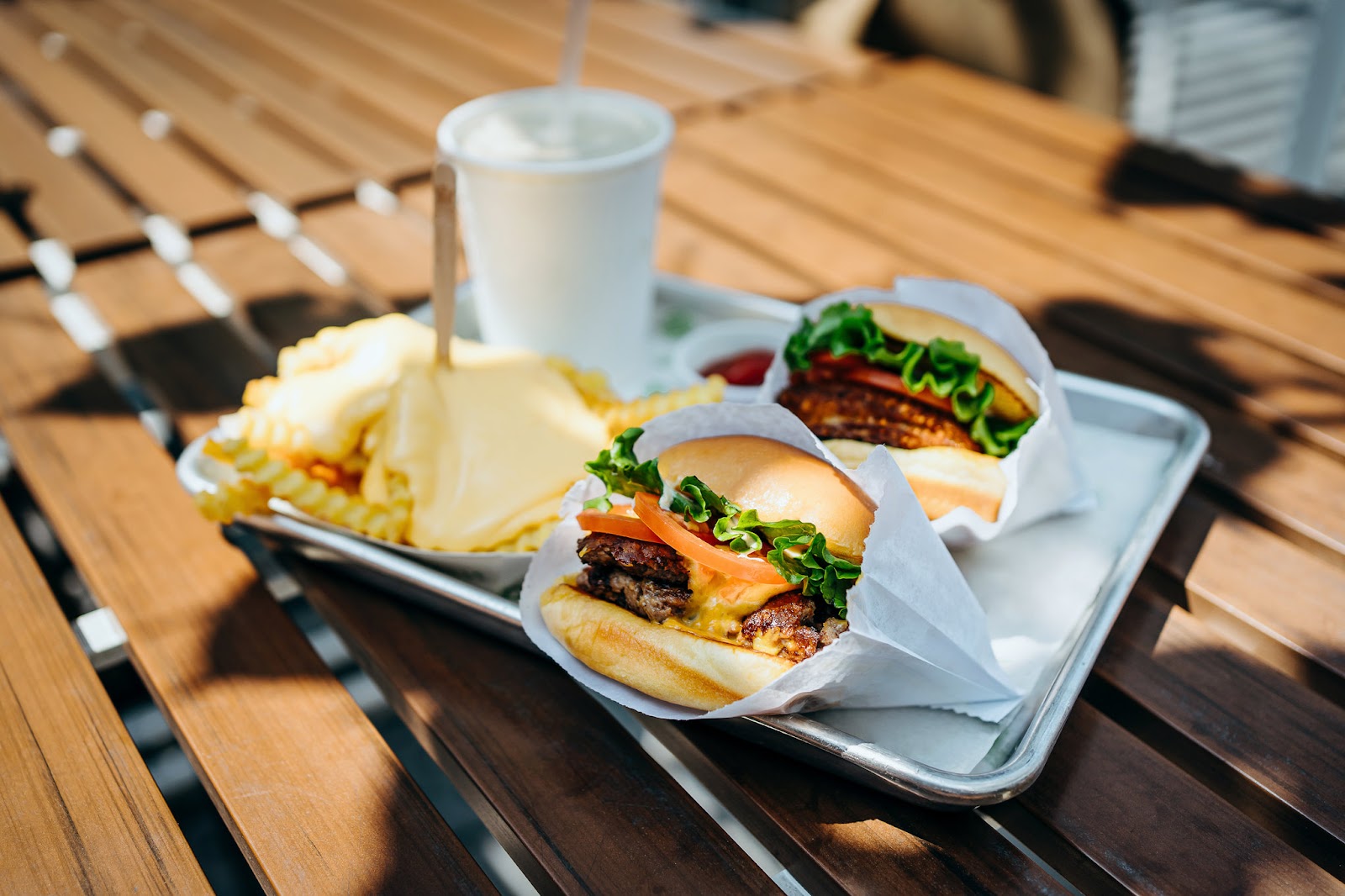 Two beef burgers with cheese and greens on paper wrapping, fries, and a drink sit on a wooden deck table in bright sunlight.