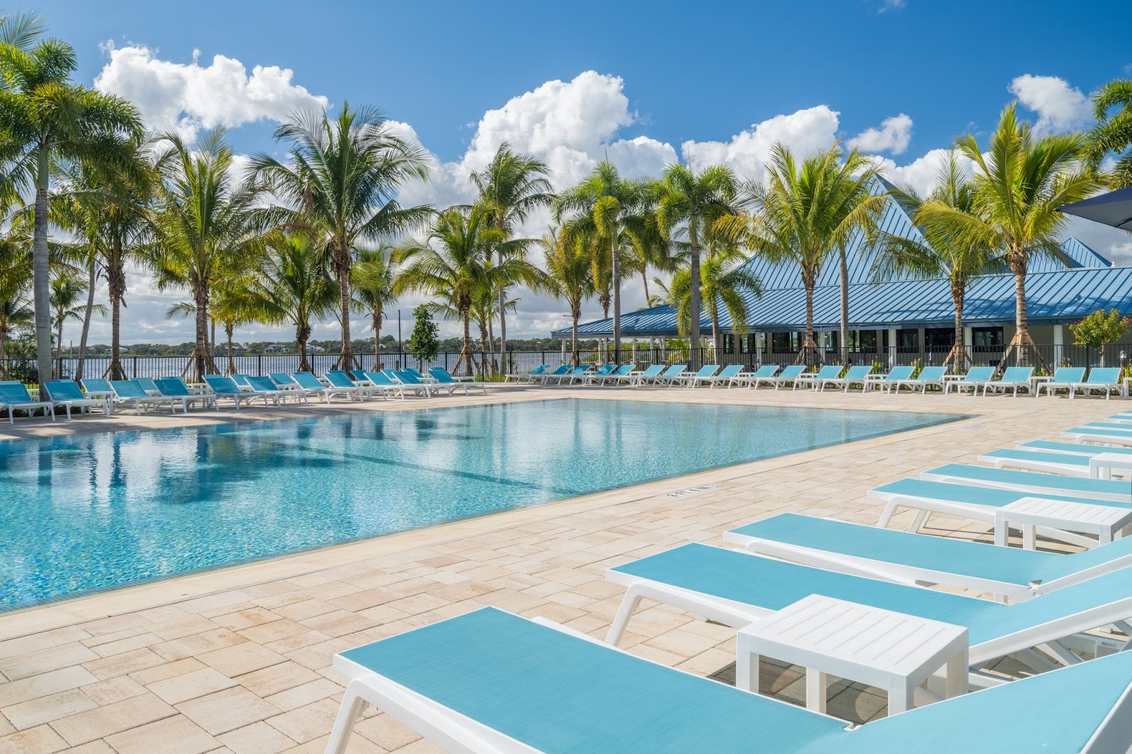 Resort pool with turquoise loungers and palm trees, overlooking a calm lake with the main building and blue metal roof visible beyond.