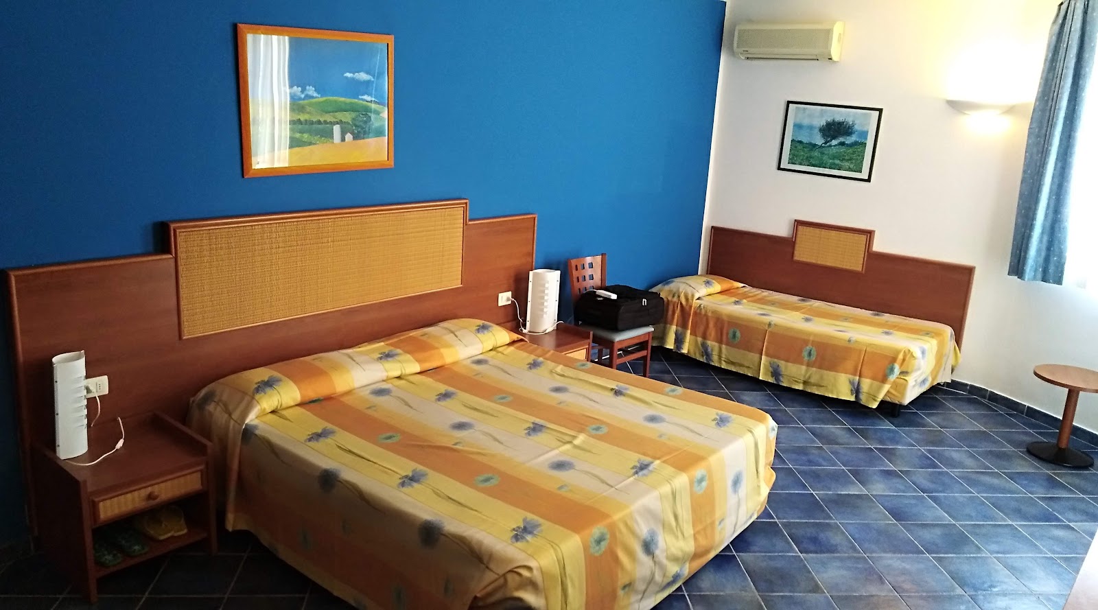 Triple guest room with yellow patterned bedspreads, wooden headboards, blue walls, and framed landscape artwork at VOI Arenella Resort.