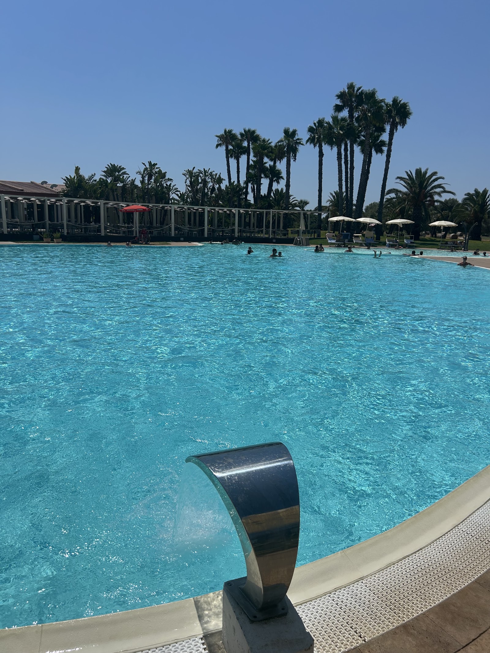 Large turquoise pool with lounge chairs, umbrellas, and palm trees at VOI Arenella Resort under clear sky.