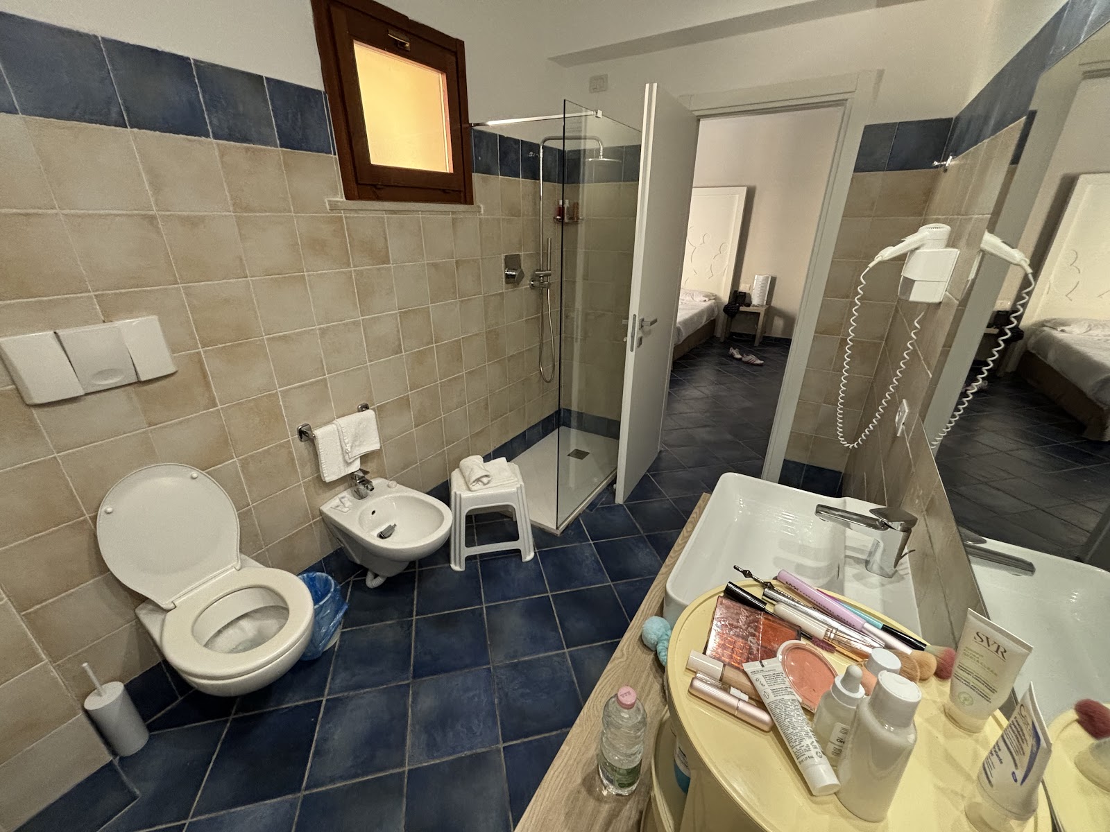 Bathroom at VOI Arenella Resort with beige and navy tile, glass shower enclosure, toilet, bidet, and vanity with toiletries.