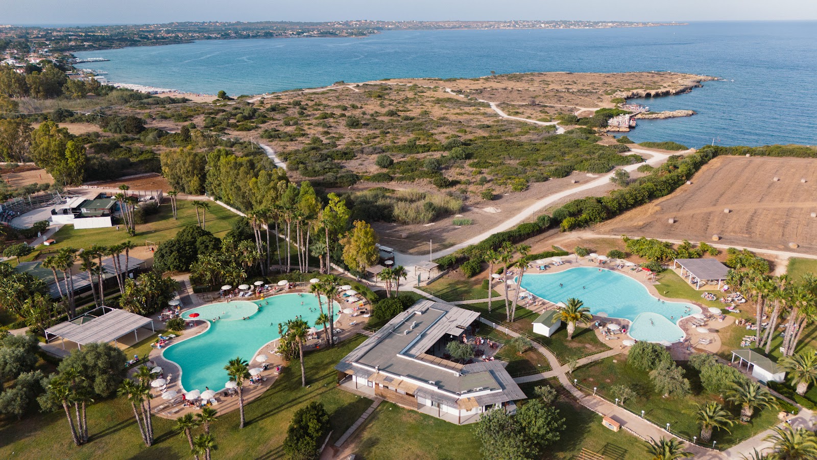 Aerial view of VOI Arenella Resort with two pools, palm trees, and Mediterranean coastline with sandy peninsula beyond.