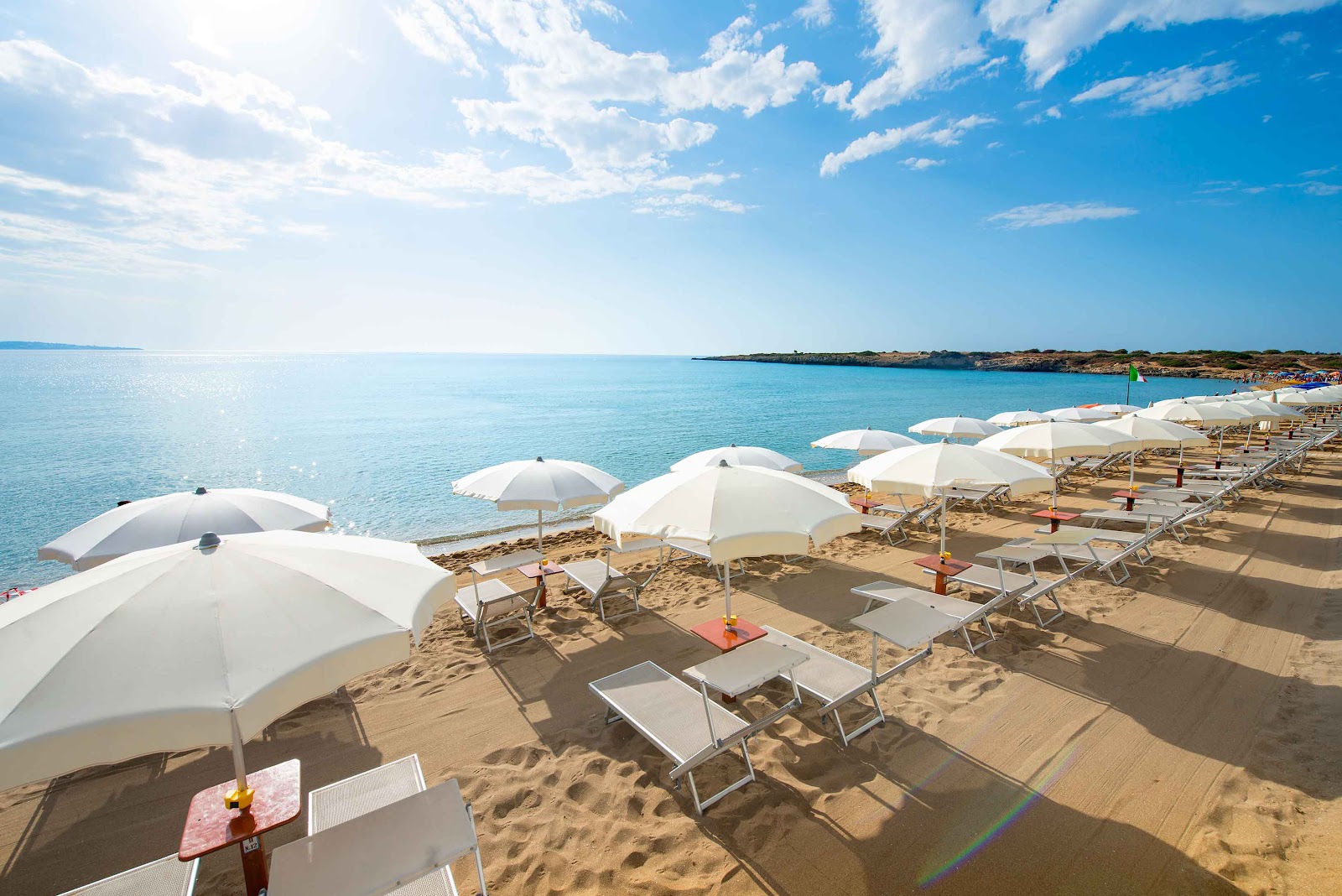 White umbrellas and loungers line the sandy beach at VOI Arenella Resort, with turquoise water and rocky coastline beyond under a partly cloudy sky.