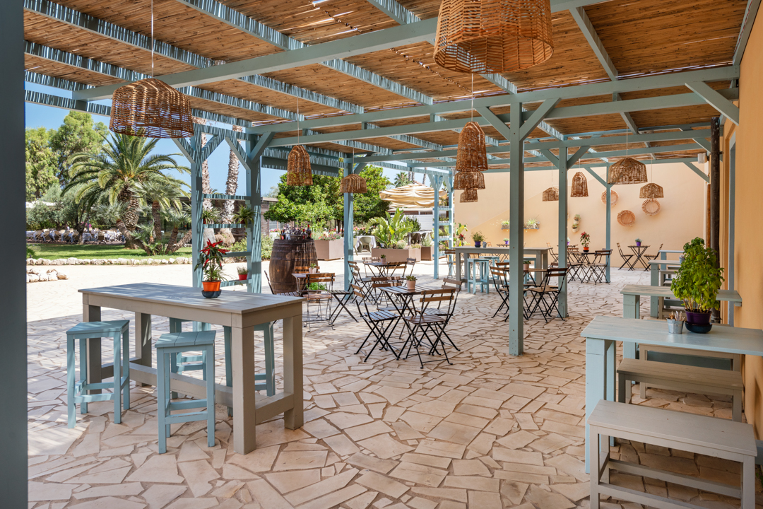 Covered terrace with wooden pergola, woven pendant lights, dining tables and chairs, and peach-colored building walls at VOI Arenella Resort.